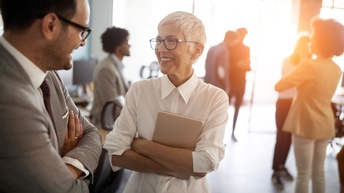 Lächelnde Person mit Brillen hält Tablet in verschränkten Armen und blickt auf andere mit ihr im Gespräch befindliche Person in Seitenansicht, im Hintergrund verschwommen weitere Personen