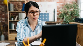 Person blickt konzentriert in einen Laptop an einem Schreibtisch, Bürosituation im Hintergrund