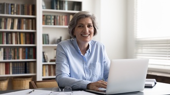 Seniorin blickt lächelnd in die Kamera und tippt dabei in ihren Laptop, daneben liegen einige Papierblätter sowie ein Stift, eine Brille und ein Notizblock, im Hintergrund stehen Bücherregale