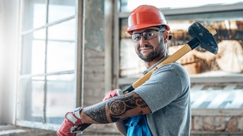 Portrait einer Person mit Schutzbrillen und rotem Helm in Arbeitslatzhose, die großen Vorschlaghammer über Schulter geschwungen hält