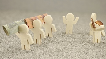 Nahaufnahme eines hellen Bodens, auf dem sechs kleine Figuren von Personen aus einem hellen Material sind. Drei Figuren tragen Geldscheine. Zwei Figuren tragen Münzen. Eine dieser Figuren hat einen nach unten geneigten Kopf