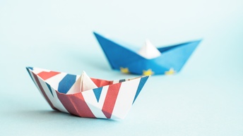 Zwei Papierschiffchen sind auf einem hellblauen Untergrund platziert, das vordere mit United Kingdom Flagge gefaltet und das hintere mit der Symbolik der Europäischen Union