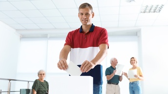 Person mit rotweißblau gestreiften T-Shirt hält Umschlag in Händen, den sie in weiße Box einwirft, im Hintergrund leicht verschwommen weitere Personen