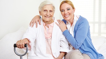 Ältere Person mit weißen Bademantel hält einen Gehstock, während eine zweite Person die Hände stützend um sie legt, beide blicken freudig in die Kamera