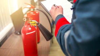 Nahaufnahme zweier Feuerlöscher, auf einem Tisch stehend. Rechts neben den Feuerlöschern steht eine Person, die in der linken Hand einen Stift hält, mit der rechten ein Blatt Papier
