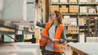 Person mit Warnweste überblickt Ware in Lagerhalle und hält ein Tablet in der Hand