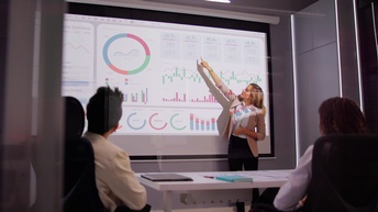 Eine Person steht vor einer großen Leinwand, auf der unterschiedliche Statistiken sind. Die Person deutet auf eine Zahl. Ihr gegenüber sitzen zwei Personen an einem Tisch, die zur Leinwand und zur Person blicken