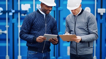 Frontalaufnahme zweier nebeneinander stehenden Personen mit weißen Helmen und Jacke. Die linke Person hält ein Tablet, die rechte ein Klemmbrett. Hinter den Personen sind blaue Container