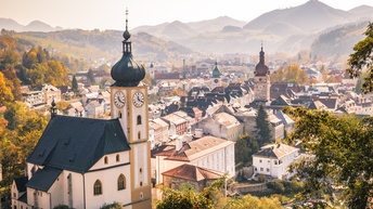 Eingebettet in eine hügelige Landschaft ist eine Ortschaft mit ein paar Kirchtürmen sowie alten Häusern. Im Vordergrund sind grüne Bäume. Im Hintergrund sind grüne Hügel
