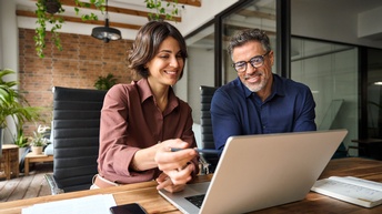 Zwei Personen sitzen an einem Tisch. Vor ihnen ist ein aufgeklappter Laptop. Beide blicken darauf. Die linke Person deutet mit einem Stift auf den Bildschirm. Neben Laptop sind Zettel und ein Notizbuch