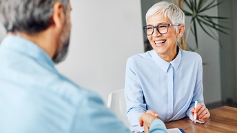 Lächelnde Person mit Brillen sitzt mit weiterer Person in Rückenansicht an Tisch und schüttelt Hand