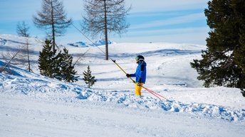 Seitliche Aufnahme einer Person in Skikleidung, Skischuhen und Ski, die sich an einer schrägen Stange festhält, die an einem Seil befestigt ist. Unter ihr und hinter ihr ist Schnee und ein blauer Himmel mit einigen Wolken