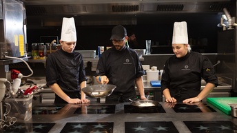 Drei Personen in Kochmontur und mit Kochhauben stehen an großem Herd mit mehreren Herdplatten, eine Person schwenkt Pfanne