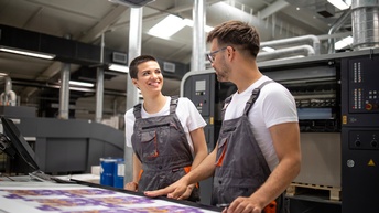 Zwei Personen in Arbeitslatzhosen stehen an Tisch mit Druckbögen und blicken einander an, im Hintergrund große Druckmaschine
