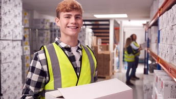 Lächelnde Person in gelber Schutzweste hält weißes Paket in Händen, im Hintergrund verschwommen weitere Personen in Gang einer Lagerhalle vor Regalen stehend