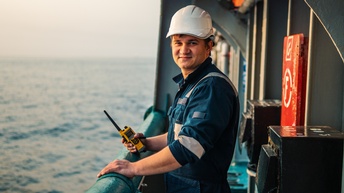 Person in Arbeitsoverall mit weißem Helm steht an Reling eines Bootes und hält Walkie Talkie in der Hand, im Hintergrund verschwommen Meer