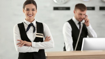 Person in Arbeitskleidung steht vor einem Empfang mit verschränkten Armen und freundlichen Blick, im Hintergrund steht eine weitere Person in Arbeitskleidung, die telefoniert und dabei auf einen Bildschirm-Monitor blickt