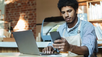 Eine Person mit Schürze sitzt an einem kleinen, runden Tisch und blickt auf ein Handy, das sie hält. Auf dem Tisch steht ein aufgeklappter Laptop. Im Hintergrund ist ein Tresen