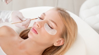 Person mit blonden langen Haaren liegt auf einer Kosmetikliege und ihre Wimpern werden gefärbt