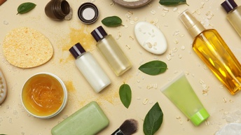 Top-Shot kleiner Fläschchen mit Flüssigkeiten, Seifen, Cremen, eines Schminkpinsels, Verschlüssen, einzelnen grünen Blättern sowie verstreuten einzelnen weißen Blütenblättern auf hellem Untergrund