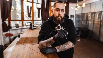 Portrait einer Person mit schwarzem Bart, Kinnpiercing, tätowierten Unterarmen und schwarzen Gummihandschuhen: Lehnt an Holztresen und hält Tätowiermaschine in einer Hand, im Hintergrund verschwommen Lichtet vor großen Fenstern mit Holzrahmen