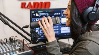 Eine Person mit Haube und Kopfhörern sitzt in einem Tonstudio und spricht in ein Mikrofon, das vor ihr hängt. Neben der Person ist ein Tisch, auf dem ein Mischpult, ein Bildschirm und eine Tastatur sind