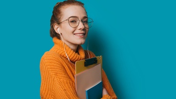Seitliche Aufnahme einer Person mit geschlossenen Haare, Brille, Kopfhörern und Heften in den Armen. Die Person trägt einen orangen Rollkragenpullover vor einer blauen Wand und lächelt
