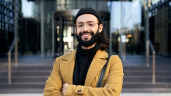 Lächelnde Person mit schulterlangen Haaren, Bart, Mütze und Brille steht mit einem gelben Mantel und verschränkten Armen vor einem modernen Gebäudeeingang aus Glas