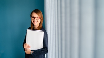 Person mit langen braunen Haaren und Brille hält Unterlagen und steht freudig vor einer blauen Wand und einer geschlossenen Fensterfront mit grauen Lamellenvorhang