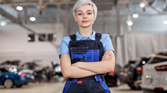 Halbporträt einer Person in einer blauen Latzhose, hellen, kurzen Haaren, Ohrringen und verschränkten Armen vor dem Bauch. Hinter der Person stehen viele Autos. Die Autos und die Person sind in einer hohen Halle