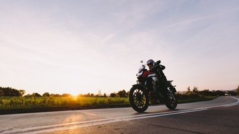 Seitliche Aufnahme einer Person auf einem Motorrad. Das Motorrad ist auf einer asphaltierten Straße. Die Person trägt einen Helm und Handschuhe. Links von der Straße ist eine grüne Wiese. Am Horizont steht die Sonne