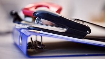 Auf einem Tisch sind Büromaterialien platziert. Im Fokus ist eine Heftklammermaschine, die auf einem Ordner liegt. Im Hintergrund sieht man verschwommen einen Locher. 