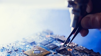 Nahaufnahme einer elektronischen Leiterplatte mit verschiedenen Mikrochips und Anschlüssen. Eine Hand hält eine Pinzette über einem kleinen elektronischen Bauteil. Mehrere Komponenten und Leiterbahnen sind auf der Leiterplatte