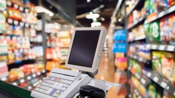 Im Vordergrund ist seitlich ein Förderband einer Kassa in einem Supermarkt. An der Kassa sind ein Bildschirm mit Tastatur sowie ein Gerät zum Scannen montiert. Im Hintergrund ist ein Gang mit Lebensmitteln befüllten Regalen.