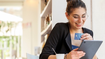 Lächelnde Person mit Brillen sitzt an Tisch, hält in einer Hand Bankomatkarte, in anderer Tablet, auf das sie blickt, vor ihr stehend weiße Tasse, im Hintergrund verschwommen Bücherregal