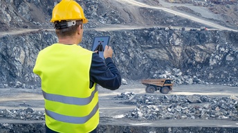 Rückenansicht einer Person mit Sicherheitsweste und Helm, die im Außenbereich eines Bergwerks stehen und ein Tablet hält. Auf dem Tablet ist die Grafik einer Straße sowie Pfeile. Im Hintergrund steht ein Fahrzeug mit einer großen Abladefläche