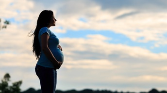 Seitliche Aufnahme einer schwangeren Person, die ihre Hand auf ihrem Bauch hat. Im Hintergrund sind grüne Bäume