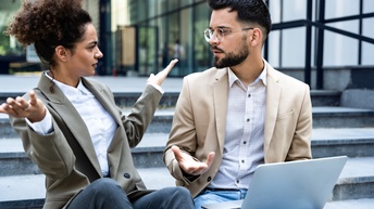 Zwei Personen in Sakkos und mit Hemden sitzen direkt nebeneinander auf einer Außentreppe und diskutieren miteinander. Die linke Person hebt die Arme. Die rechte Person hat auf dem Schoß einen aufgeklappten Laptop