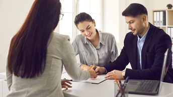 Rückenansicht einer Person in einem hellgrauen Sakko, die an einem Tisch sitzt. Der Person gegenüber sitzen zwei Personen. Zwischen ihnen liegt ein Klemmbrett mit Zetteln, auf denen die rechte Person unterschreibt