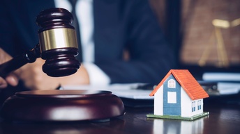 Auf einem Tisch steht ein Modell eines kleinen Hauses. Daneben ist ein Richterhammer