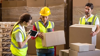 Drei Personen mit gelben Schutzwesten inspizieren Kartonagen, eine Person trägt gelben Helm am Kopf, eine andere bedient ein Tablet