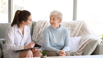 Person in weißer Arbeitskleidung sitzt bei einer betagten Person auf einer Couch und hält ein Klemmbrett sowie einen Stift in der Hand, im Hintergrund zeigt sich ein Wohnraum