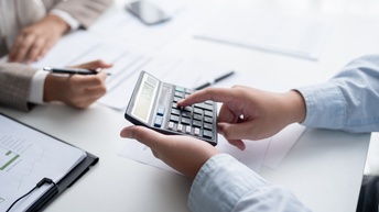Zwei Personen sitzen sich an einem Tisch gegenüber. Die rechte Person tippt auf einem Taschenrechner. Die linke Person notiert mit einem Stift etwas auf einem Zettel