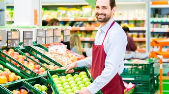 Seitliches Halbporträt einer Person mit roter Schürze, die mit beiden Händen eine niedrige, grüne Kiste hält, in der grünes Obst liegt. Links sind weitere dieser Kisten, befüllt mit unterschiedlichem Obst. Im Hintergrund sind Regale mit Lebensmitteln