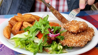 Nahaufnahme eines Schnitzels mit Sesam, Kartoffelspalten, Salt mit Gurken und Tomate sowie zweier Saucen auf einem Teller. Eine Person schneidet das Schnitzel<br />
