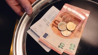 Nahaufnahme eines runden, kleinen Metalltellers, auf dem eine Rechnung, zwei zehn Euroscheine, zwei zwei Euromünzen und eine fünfzig Centmünze liegen. Das Tablett wird von einer Hand einer Person gehalten