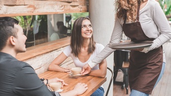 Zwei Personen sitzen sich an einem Tisch im Außenbereich eines Lokals gegenüber. Vor eine der Personen stellt eine Person mit Schürze und Tablett, die einen Kaffee vor sie stellt