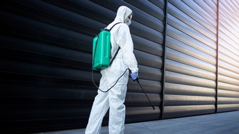 Person in Rückenansicht in weißem Schutzanzug mit Schutzbrille, Atemschutz und Handschuhen trägt türkisen Container verbunden mit Sprühstab am Rücken, vor Lamellenwand stehend