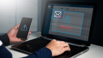 Die Hände einer Person sind auf der Tastatur eines aufgeklappten Laptops. Auf dem Bildschirm ist ein Rufzeichen in einem Dreieck, unter dem Warnung steht. Die Person hält ein Handy, auf dem ebenfalls ein Rufzeichen in einem Dreieck ist