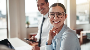 Person mit geschlossenen dunklen Haaren und Brille, sowie gestreifter Bluse blickt freudig in die Kamera während eine Person mit Hemd und Krawatte hinter ihr steht und Smartphone in der Hand hält